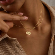Close-up of a person wearing a 14k gold-filled necklace with a 'M' pendant, with a neutral background.